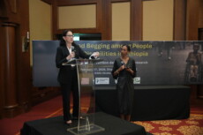 Two women on a stage at a conference: one speaks at a clear podium, the other stands beside holding a microphone, with a large banner in the background.