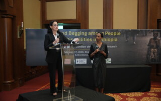 Annabel and Beza – Population Council Two women on a stage at a conference: one speaks at a clear podium, the other stands beside holding a microphone, with a large banner in the background.