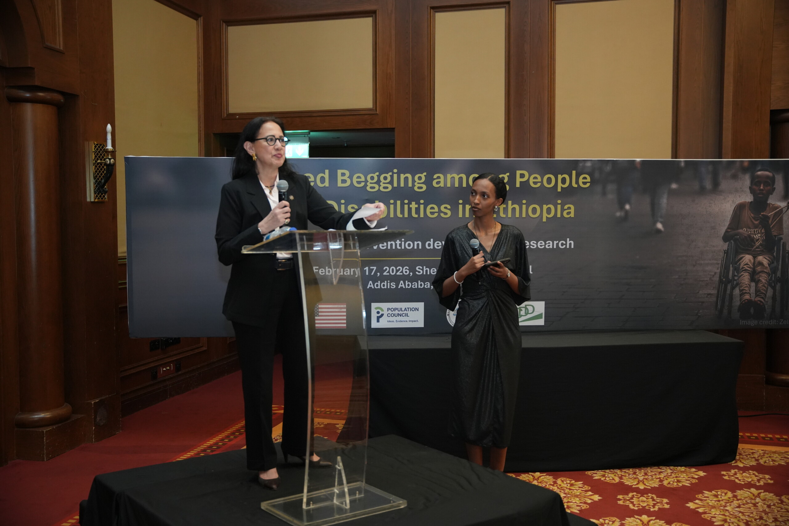 Two women on a stage at a conference: one speaks at a clear podium, the other stands beside holding a microphone, with a large banner in the background.