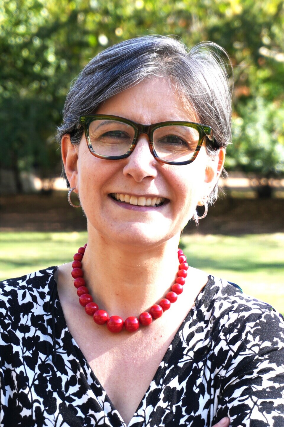 Dr. Tiziana Leone, smiling woman with short gray hair and glasses in a sunlit park backdrop